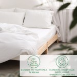 Prześcieradło satynowe z gumką w kolorze białym, rozłożone na łóżku z drewnianą ramą. Widoczne dwa białe poduszki. Tło jasne, z firankami i skromnymi roślinami, tworzy przytulną atmosferę. Szwy i faktura materiału nie są widoczne.