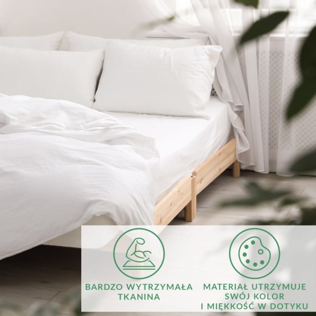 Prześcieradło satynowe z gumką w kolorze białym, rozłożone na łóżku z drewnianą ramą. Widoczne dwa białe poduszki. Tło jasne, z firankami i skromnymi roślinami, tworzy przytulną atmosferę. Szwy i faktura materiału nie są widoczne.