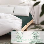 Prześcieradło satynowe z gumką w kolorze butelkowej zieleni, rozłożone na łóżku z drewnianą ramą. Na poduszce widoczne białe poszewki. Tło neutralne z zasłonami, co podkreśla kolor materiału. Wymiary 200x220 cm, idealne do dużych łóżek.