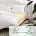 Prześcieradło satynowe z gumką w kolorze kremowym o wymiarach 200x220 cm, rozłożone na łóżku z białą pościelą. Widoczne poduszki w kolorze białym.