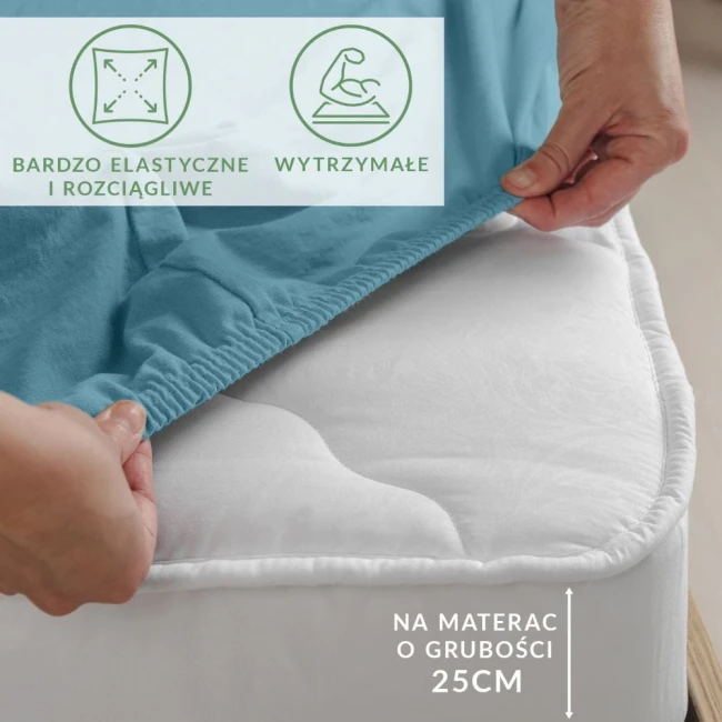 Prześcieradło jersey z elastanem w kolorze błękitnym, z gumką, na materac o grubości 25 cm, przedstawione w zbliżeniu, z ręką zakładającą je na materac. Tło neutralne, podkreślające materiał. Widoczne napisy informujące o elastyczności i wytrzymałości.