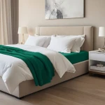 Prześcieradło jersey z elastanem w kolorze zielonym, rozmiar 160x200, leżące na łóżku z jasnym, eleganckim zagłówkiem. W tle widoczne białe poduszki i dekoracyjne akcesoria na szafce nocnej, na drewnianej podłodze.