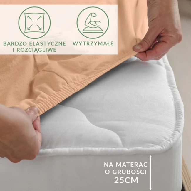 Prześcieradło jersey z elastanem w kolorze różowym, rozłożone na materacu o grubości 25 cm. Widoczne elastyczne brzegi, które zapewniają idealne dopasowanie. Ujęcie z boku, w ręce trzymane przy krawędzi, pokazuje materiał leżący na białym materacu.
