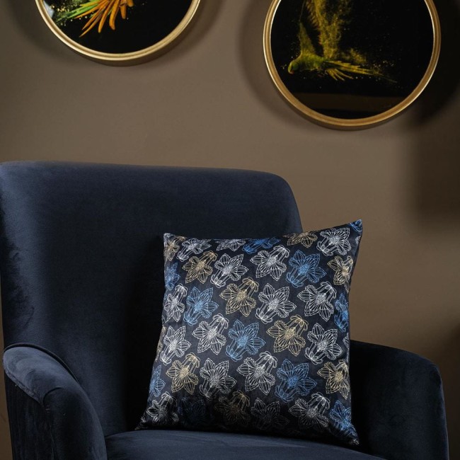 Poszewka dekoracyjna 43x43 w kolorze granatowym z kwiatowym wzorem. Zbliżenie na poszewkę leżącą na ciemnoniebieskim fotelu. Wzór przedstawia białe, niebieskie i złote kwiaty, wykonana z gładkiego materiału. Tło w odcieniach brązu.