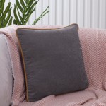 Poszewka dekoracyjna 40x40 w kolorze szarym z lamówką w odcieniu brązowym, leżąca na różowej narzucie, w aranżacji wnętrza. Kąt ujęcia frontalny, pokazujący prostokątny kształt i teksturę materiału. Tło jasne, z elementami roślinnymi.