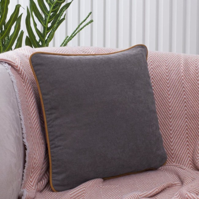 Poszewka dekoracyjna 40x40 w kolorze szarym z lamówką w odcieniu brązowym, leżąca na różowej narzucie, w aranżacji wnętrza. Kąt ujęcia frontalny, pokazujący prostokątny kształt i teksturę materiału. Tło jasne, z elementami roślinnymi.