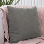 Poszewka dekoracyjna 40x40 w kolorze szarym, gładka, umieszczona na sofie. Produkt leży na jasnym kocyku w wzór chevron. Zachowuje prostokątny kształt, bez widocznych napisów ani logo. Tło jest jednolite, co podkreśla kolor poszewki.