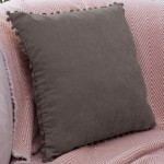 Poszewka dekoracyjna 45x45 szara, wykonana z miękkiego materiału, z dekoracyjnymi pomponami wzdłuż krawędzi. Ułożona leżąco na różowym tle, które posiada wzór w zygzaki. Zbliżenie na teksturę tkaniny, subtelnie podkreślającą kontrast z tłem.