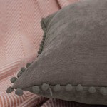 Poszewka dekoracyjna 45x45 cm w szarym kolorze, z miękkiego materiału, z pomponami przy brzegach. Ujęcie w zbliżeniu na detal faktury i zamka. Tło w pastelowym odcieniu z wyraźnym wzorem, co podkreśla produkt.