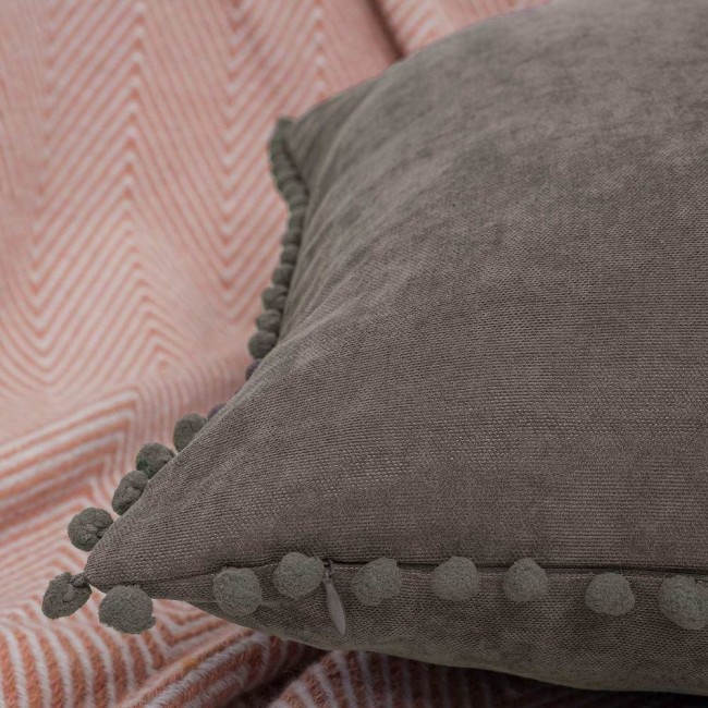 Poszewka dekoracyjna 45x45 cm w szarym kolorze, z miękkiego materiału, z pomponami przy brzegach. Ujęcie w zbliżeniu na detal faktury i zamka. Tło w pastelowym odcieniu z wyraźnym wzorem, co podkreśla produkt.
