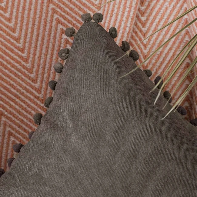 Poszewka dekoracyjna 45x45 cm w szarym kolorze, z pomponami przy krawędzi. Ujęcie z boku, w zbliżeniu, ukazujące fakturę materiału. Tło w geometryczny wzór w odcieniach różu, tworzy kontrast. Produkt leży na poziomej powierzchni obok rośliny.