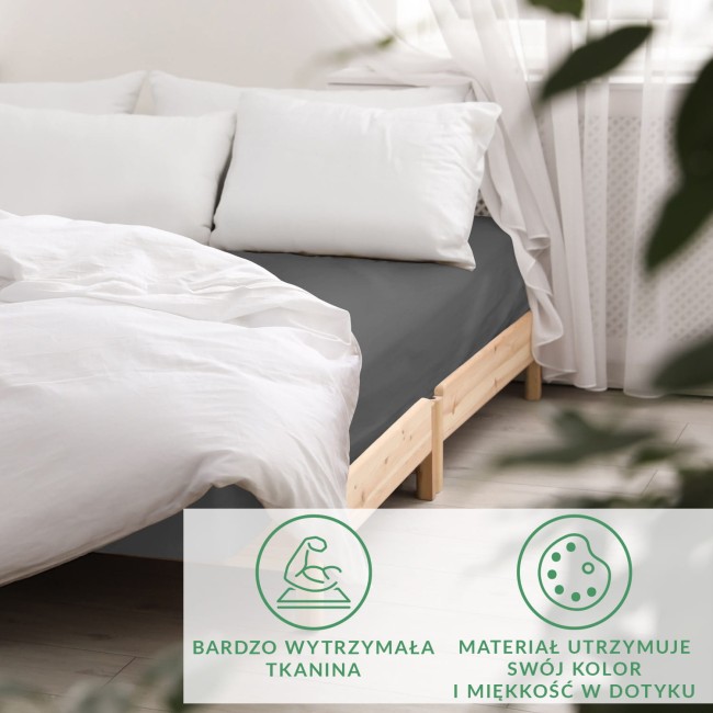 Prześcieradło satynowe bez gumki w kolorze szarym, leżące na łóżku, w zestawieniu z białymi poduszkami. Produkt widoczny z boku, eksponujący teksturę materiału. W tle jasne wnętrze z oknem i firaną, podkreślające elegancję aranżacji.