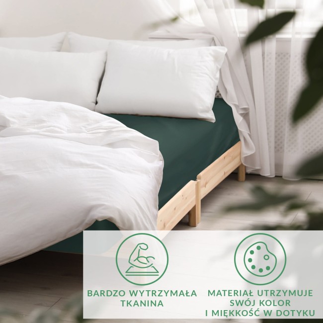 Prześcieradło satynowe bez gumki w kolorze butelkowej zieleni, rozłożone na łóżku z jasnym drewnianym stelażem. Białe poduszki znajdują się na górze. Tło w jasnych, naturalnych kolorach, co podkreśla głęboki odcień zieleni materiału.