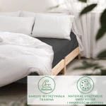 Prześcieradło satynowe bez gumki w kolorze czarnym o wymiarach 200x220 cm, leżące na łóżku z białymi poduszkami. Kąt ujęcia z boku, na jednolitym tle. Materiał gładki, błyszczący, z przyjemną w dotyku fakturą. W tle widoczne białe firany.