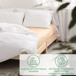 Prześcieradło satynowe bez gumki w kolorze jasnym beżu, rozłożone na łóżku, wyeksponowane z boku. Widoczne są białe poduszki i lekka, jasna pościel. Tło prostego, jasnego wnętrza. Produkt uwydatnia miękkość materiału.