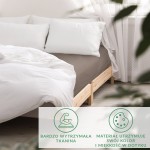 Prześcieradło satynowe bez gumki w kolorze Sepia, rozmiar 200x220 cm, leżące na łóżku z białymi poduszkami. Widoczne drewniane nogi łóżka oraz jasne tło z zasłonami. Tkanina charakteryzuje się gładką, błyszczącą fakturą.