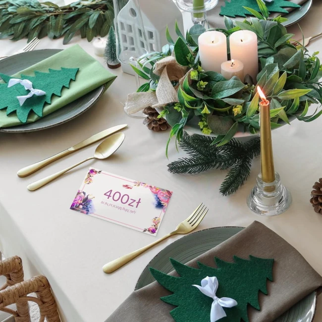 Karta podarunkowa o wartości 400 zł leży na beżowym obrusie, otoczona dekoracyjnymi elementami stołowymi, takimi jak sztućce złotego koloru, zielone serwetki w kształcie choinek oraz ozdobna świeczka. W tle widoczne rośliny dekoracyjne.