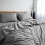 Pościel satyna bawełniana w kolorze popielatym, o wymiarach 140x200 cm, leżąca na łóżku. Widoczne dwa poduszki w tym samym kolorze. Całość w prezentacji minimalistycznej, na tle jasnej ściany oraz drewnianego zagłówka.