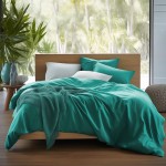 Pościel satyna bawełniana w kolorze ciemnego turkusu o wymiarach 180x200. Widok z boku, produkt rozłożony na łóżku, otoczony białymi poduszkami i dekoracyjnym kocem w odcieniu turkusu. Otoczenie w jasnym wnętrzu z roślinnością w tle.