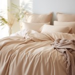 Poszewka satynowa 40x40 w kolorze jasnego beżu, leżąca na pościeli, z widoczną gładką fakturą. W tle poduszki w tym samym odcieniu oraz dodatkowy koc w kolorze beżowym. Scena w naturalnym świetle, tworzy przytulny nastrój.