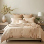 Poszewka satynowa 70x80 w jasnym beżu, leżąca na łóżku z rozłożonymi poduszkami. W tle widoczna roślina w wazonie oraz lampka nocna. Materiał gładki, o delikatnym połysku, dobrze komponuje się z neutralnym wnętrzem.