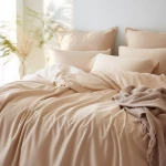 Poszewka satynowa 70x80 w kolorze jasnego beżu leżąca na łóżku, z widoczną gładką fakturą. Obok kilka poduszek w tym samym odcieniu oraz lekki koc w odcieniu taupe. Całość na tle neutralnej ściany, tworząc stonowaną aranżację.