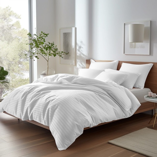 Poszewka satyna bawełniana 70x80 w kolorze białym, umieszczona na łóżku z jasnym drewnianym stelażem, obok białych poduszek w pasy. Tło przedstawia jasne wnętrze z oknem oraz rośliną doniczkową. Tekstura materiału gładka.