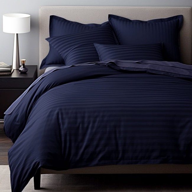 Poszewka satynowa bawełniana 40x40 Cizgili w kolorze ciemnym granacie, z widoczną fakturą w poziome paski, leży na łóżku. W zestawie kilka poduszek o różnych rozmiarach i kształtach w tym samym odcieniu, na tle jasnej ściany.