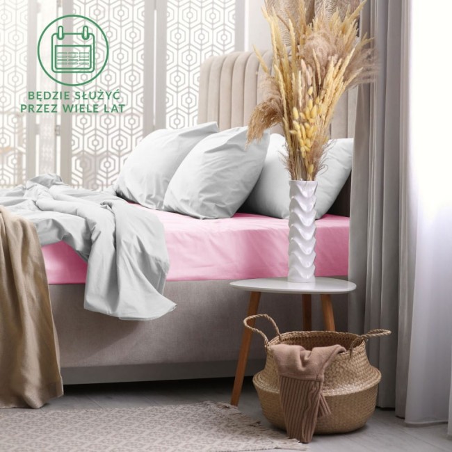Prześcieradło frotte z gumką 60x120 w kolorze jasnego różu, rozłożone na łóżku, widoczne z boku. W otoczeniu szarej pościeli i jasnych poduszek, obok stolika z białym wazonem i dekoracjami. Tło neutralne, jasne, tworzy przytulną aranżację sypialni.