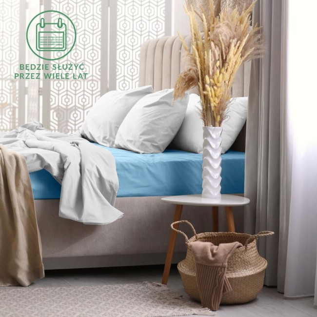 Prześcieradło frotte z gumką 60x120 w kolorze niebieskim, rozłożone na łóżku, z widocznym jasnobrązowym kocem. Otoczenie zawiera białe poduszki, dekoracyjne kwiaty w wazonie oraz stół z plecionym koszem. Tło stanowią zasłony i wzorzysty panel.