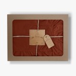 Pościel bawełniana z falbanką w kolorze ceglanym, o wymiarach 160x200, zapakowana w solidne, ozdobne opakowanie z tektury. Widoczne detale to subtelne falbanki oraz ręczna metka z logo producenta. Produkt leży na tle białego tła.