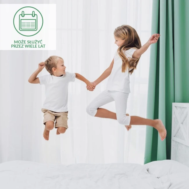 Ochraniacz na materac 70x140 Robson w kolorze białym leży na łóżku. Na zdjęciu dzieci skaczą na materacu, trzymając się za ręce. Tło to jasne zasłony oraz zielone firany, co tworzy przyjemną aranżację wnętrza.