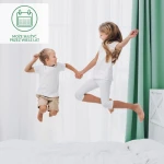 Ochraniacz na materac 100x200 Robson w kolorze białym, zbliżenie na dzieci skaczące na łóżku z białą narzutą. Białe, gładkie tło z delikatnymi zasłonami oraz zielonymi firanami, dzieci w ubraniach w jasnych kolorach, trzymają się za ręce.