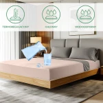 Ochronne prześcieradło z gumką 90x200 SMARTCEL w kolorze beżowym, leżące na łóżku z szarym zagłówkiem. Widać szklankę wody i wylewający się napój. Widoczne oznaczenia funkcji: termoregulacyjny, chłonny, wodoodporny. Tło w neutralnych odcieniach.