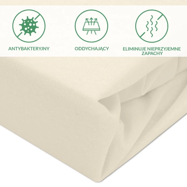 Ochronne prześcieradło z gumką w kolorze kremowym o wymiarach 60x120 cm, leżące złożone. Materiał o teksturze bawełny, widoczne zakładki. Elementy na górze przedstawiają właściwości: antybakteryjny, oddychający, eliminuje nieprzyjemne zapachy.