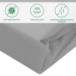 Ochronne prześcieradło z gumką w szarym kolorze, złożone, ukazane z boku. Materiał ma gładką teksturę. W górnej części widoczne grafiki przedstawiające właściwości: antybakteryjność, oddychalność oraz eliminację nieprzyjemnych zapachów.