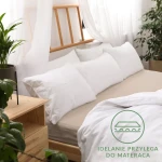 Beżowe prześcieradło jersey z gumką w rozmiarze 80x180, rozłożone na łóżku, dobrze przylegające do materaca. W tle widoczne białe poduszki i drewniana rama łóżka. Delikatne tło z roślinnością i zasłonami.