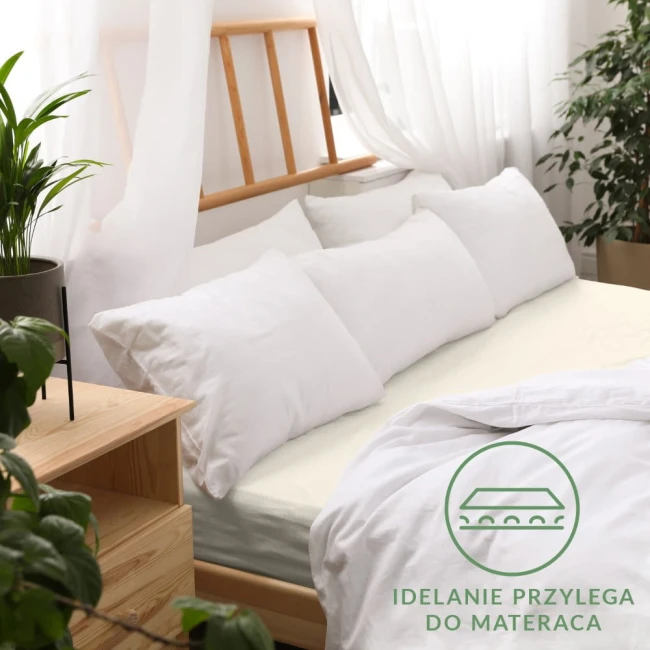 Prześcieradło jersey z gumką o wymiarach 80x180 w kolorze ecru, rozłożone na łóżku. Ujęcie z boku, prezentujące gładką fakturę materiału oraz gumkę przylegającą do materaca. Tło w jasnym wnętrzu z roślinnością i drewnianymi meblami.