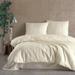 Pościel satyna bawełniana 160x200 w kolorze ecru, z geometrycznym wzorem. Złożona na łóżku, z widocznymi poduszkami. Tło neutralne, betonowe, z roślinnością w donicy i lampą biurkową w aranżacji. Gładka tekstura materiału.