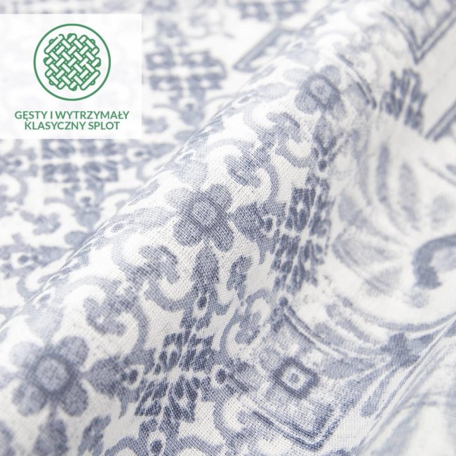 Pościel bawełniana Cottonlove Exclusive Pava w kolorze białym z niebieskim ornamentem, złożona w pół, ukazująca teksturę materiału. Wzór floralny z delikatnymi zdobieniami. Widoczne logo i napis, tylko w dolnej części zdjęcia, na jasnym tle.