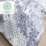Pościel bawełniana 200x220 Cottonlove Exclusive Pava w kolorze białym z niebieskim ornamentem. Ułożona na stole w zbliżeniu, ukazuje różnorodne wzory, w tym liście i geometryczne motywy, z widoczną teksturą materiału.