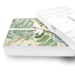 Pościel satyna bawełniana 220x200 w zielonym kolorze z dekoracyjnymi liśćmi, zapakowana w białe pudełko z logo Luxury. Widoczna wstążka na opakowaniu. Perspektywa z góry, ukazująca produkt leżący w otwartym pudełku.