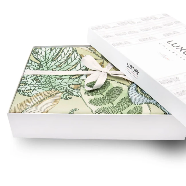 Pościel satyna bawełniana 220x200 w zielonym kolorze z dekoracyjnymi liśćmi, zapakowana w białe pudełko z logo Luxury. Widoczna wstążka na opakowaniu. Perspektywa z góry, ukazująca produkt leżący w otwartym pudełku.