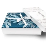 Pościel satyna bawełniana Luxury Premium Irene w rozmiarze 220x200, zapakowana w eleganckie opakowanie w kolorze niebieskim z delikatnym wzorem liści. Opakowanie zamknięte białą wstążką, widoczny napis LUXURY. Górna część opakowania lekko uniesiona.