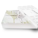 Pościel satyna bawełniana 220x200 Luxury Premium Palmiro w zielone dekoracyjne liście, zapakowana w eleganckie pudełko, z białą kokardą. Widoczny nadruk liści palmy na tkaninie w białym tle, opakowanie leży na białej powierzchni.