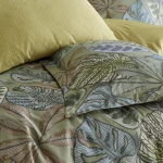 Pościel satynowa 220x200 cm w kolorze zielonym z dekoracyjnym wzorem liści. Złożone poduszki w jasnym odcieniu żółtym; faktura gładka, błyszcząca. Ujęcie z bliska, ukazujące detale wzoru i tkaniny, na neutralnym tle.