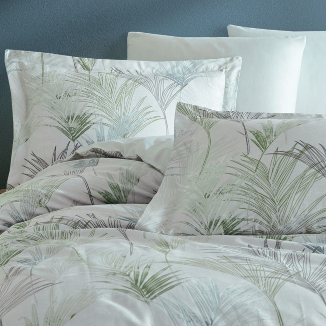 Pościel satyna bawełniana 220x200 Luxury Premium Palmiro w zielone liście, rozłożona na łóżku, z poduszkami w tym samym wzorze oraz białymi poduszkami w tle. Tło ściany w chłodnym odcieniu. Wzór liściowy subtelny, z dominującym zielonym i szarym kolorem.
