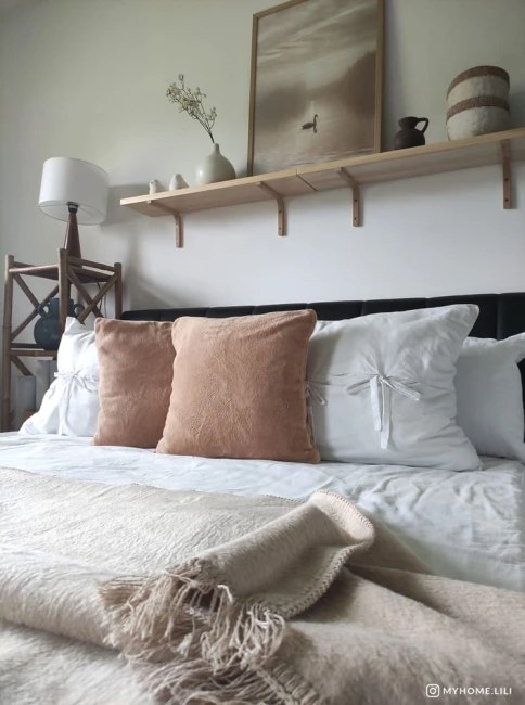Koc bawełniany w jasnym beżu o wymiarach 200x220 cm z frędzlami leży na łóżku. W tle widoczne poduszki białe i beżowe oraz elementy dekoracyjne na półce, tworzące przytulną aranżację sypialni.