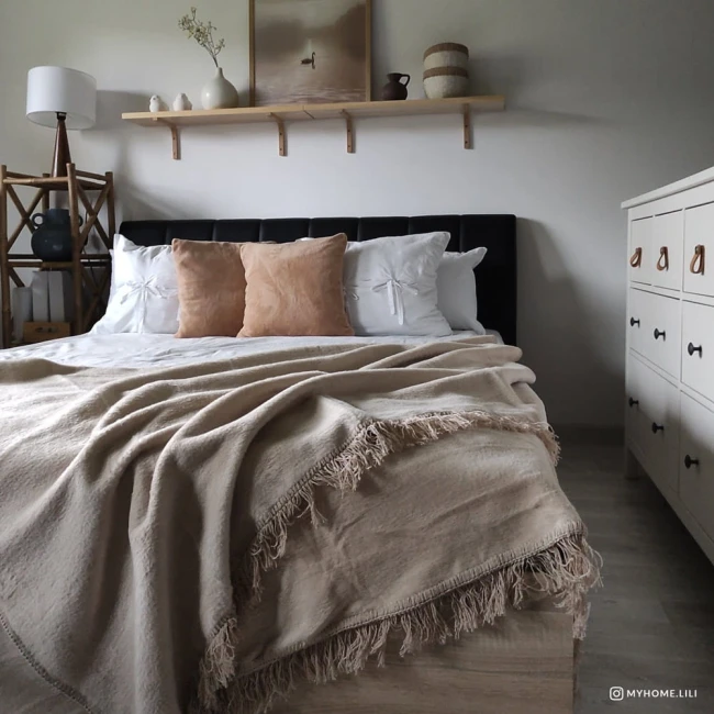 Koc bawełniany 200x220 w jasnym beżu z dekoracyjnymi frędzlami, leżący na łóżku z poduszkami w odcieniach białego i brązowego. Widok z boku, w aranżacji sypialni z jasnym tłem i drewnianymi elementami.