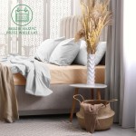 Prześcieradło frotte z gumką 200x220 w kolorze róż perłowym, rozłożone na materacu, z miękką narzutą w odcieniach beżu. W tle widoczne poduszki w szarym kolorze oraz kwiaty w białej wazonie, na stoliku. Tło jest jasne, z delikatnym wzorem.